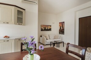 Ein Essbereich mit einem Holztisch, einer Vase mit violetten Blumen und einem Teller mit Essen. Im Hintergrund befinden sich eine Couch und ein Couchtisch.