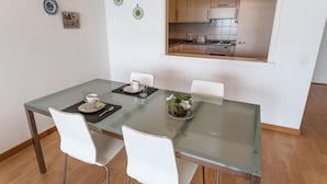 Uma área de refeições moderna com uma mesa de vidro, cadeiras brancas e vista para uma cozinha através de uma porta aberta.