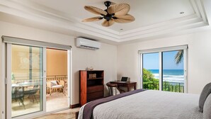 A bedroom with a bed, a desk, a chair, a bookshelf, and a sliding glass door leading to a balcony with a view of the ocean.
