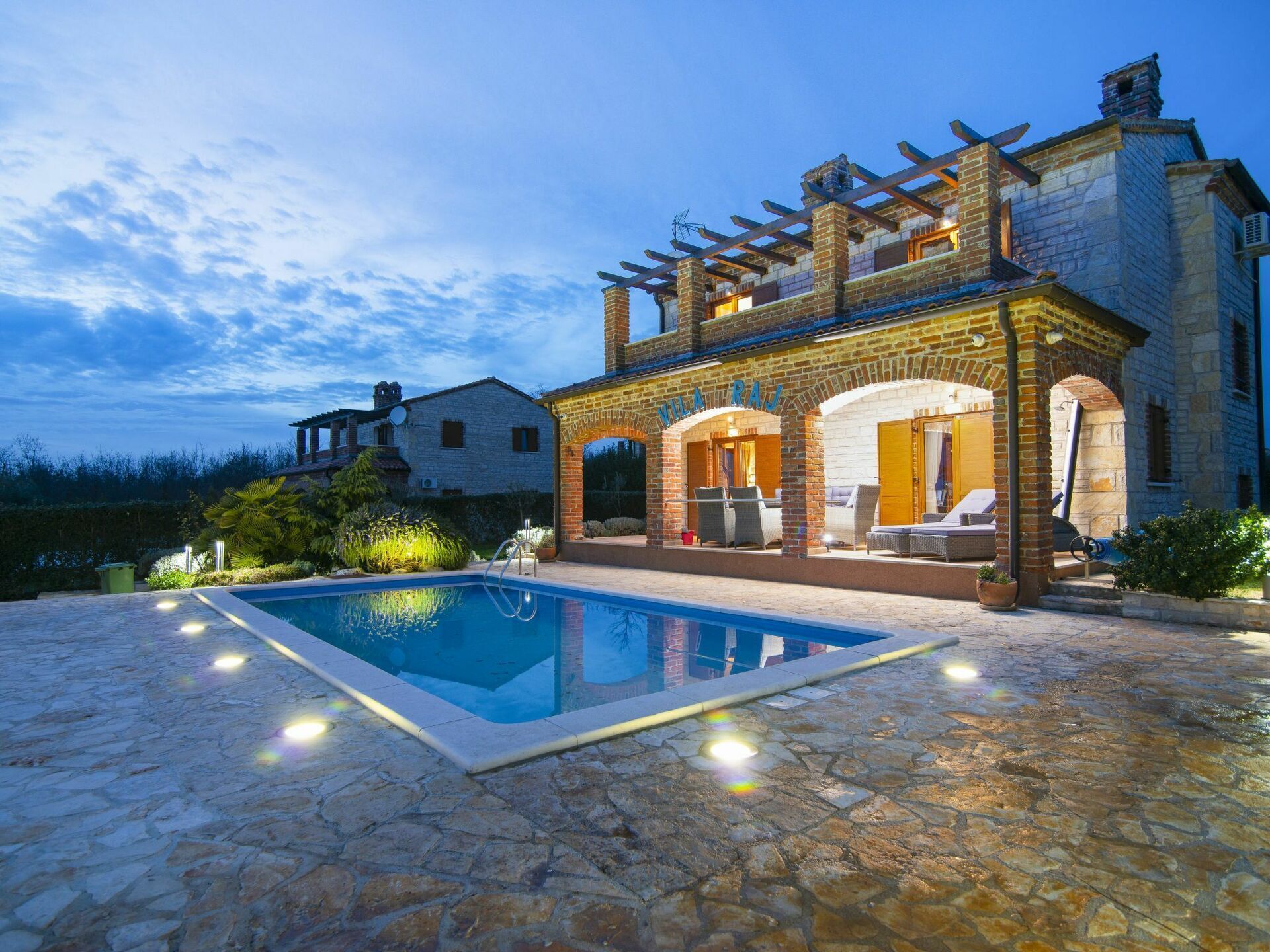 Uma piscina de pedra com pérgola e espreguiçadeiras, rodeada por um terraço de pedra.