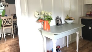 A kitchen with a white table, a vase with flowers, a framed photo, and a bowl of fruit.