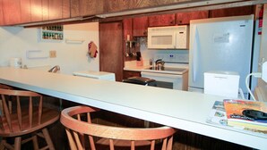 Una cocina con armarios de madera, una encimera blanca y un refrigerador. Hay dos sillas de madera y una revista sobre la encimera.