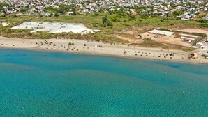Vue aérienne d’une zone côtière avec une plage de sable, des eaux turquoise et limpides, et une zone urbaine densément peuplée en arrière-plan.