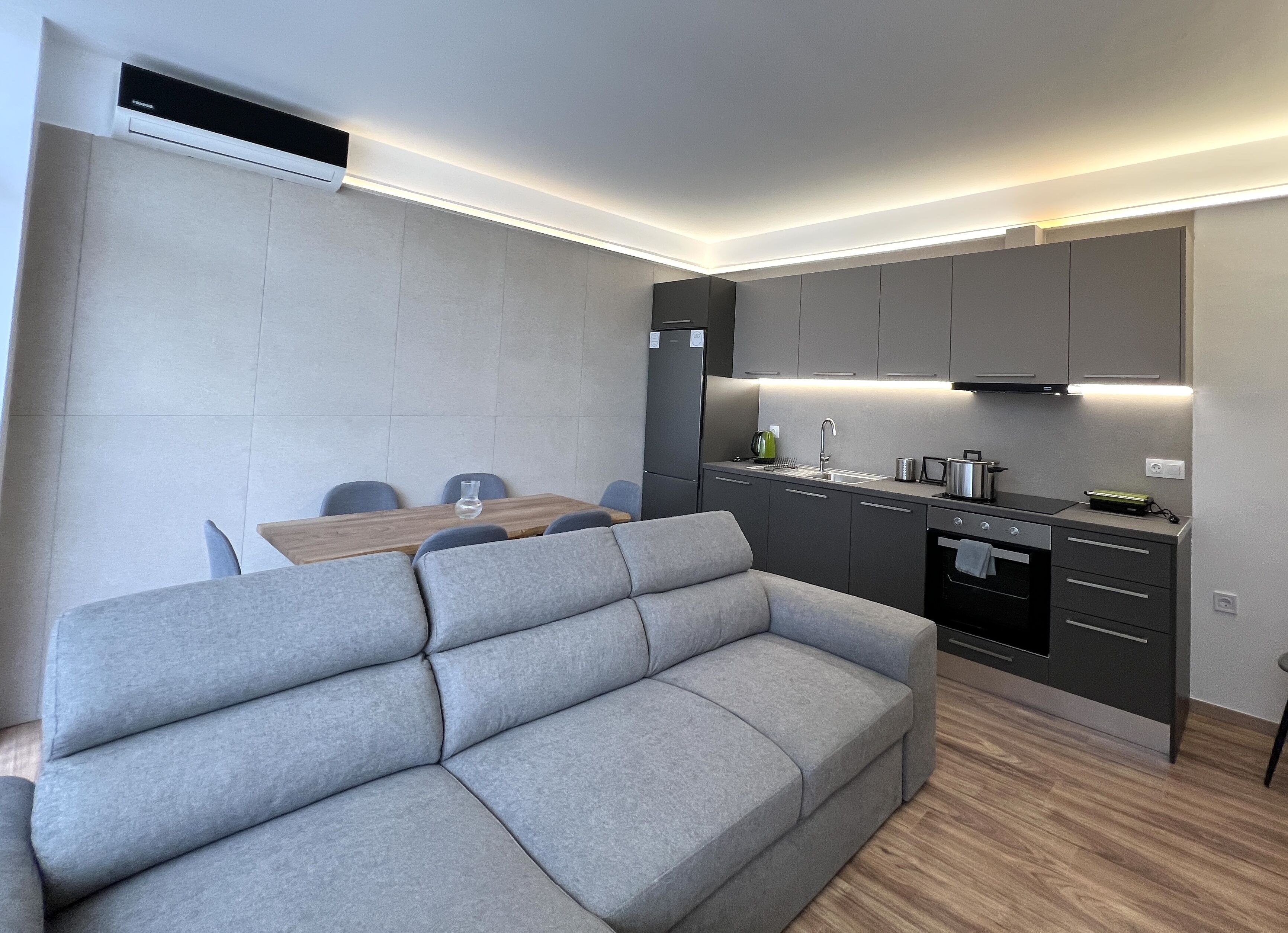 Une cuisine moderne avec des meubles gris, un four encastré et un coin repas comprenant un canapé gris et une table en bois.