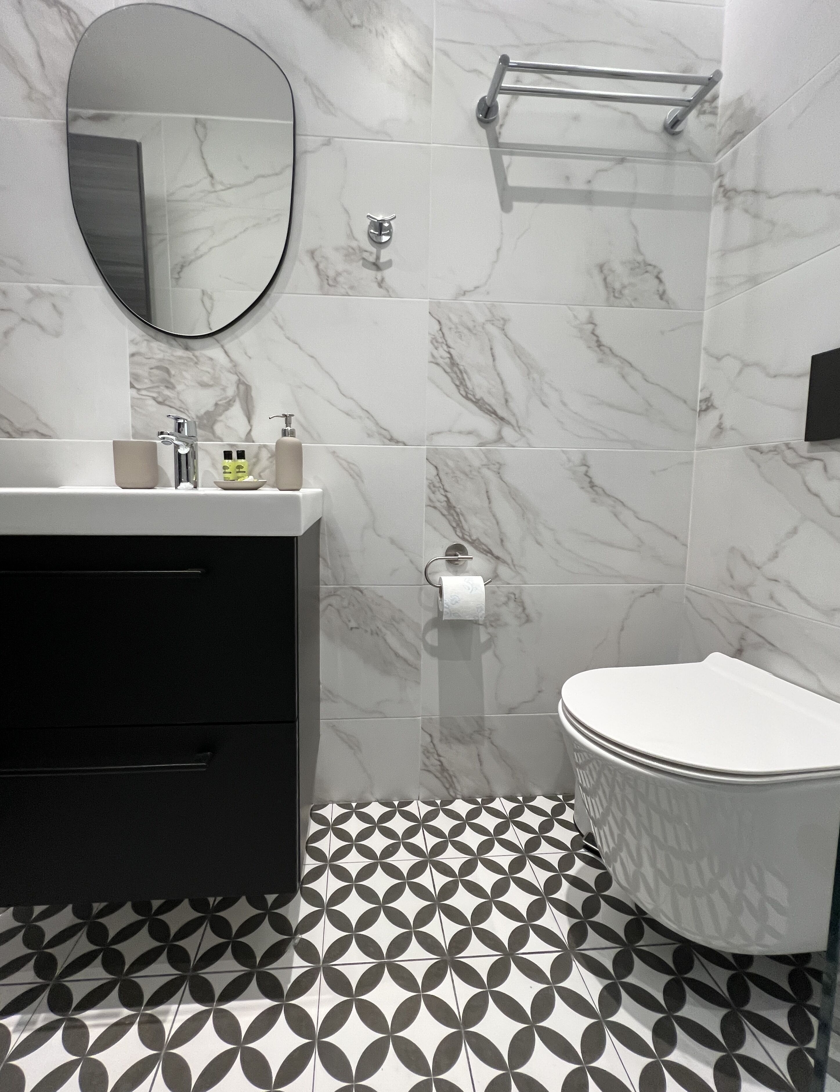 Un baño moderno con salpicadero de mármol, suelo a cuadrícula blanco y negro, espejo redondo, inodoro y tocador con cajones y dispensador de jabón.