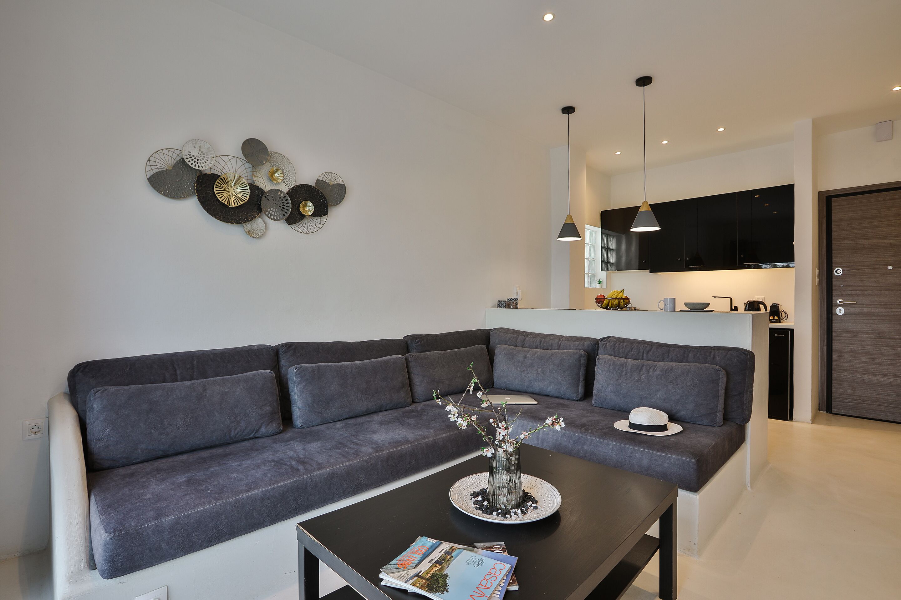 A modern living room with a grey sofa, a coffee table with a vase and magazines, and a kitchen in the background.