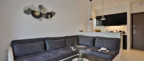 A modern living room with a grey sofa, a coffee table with a vase and magazines, and a kitchen in the background.