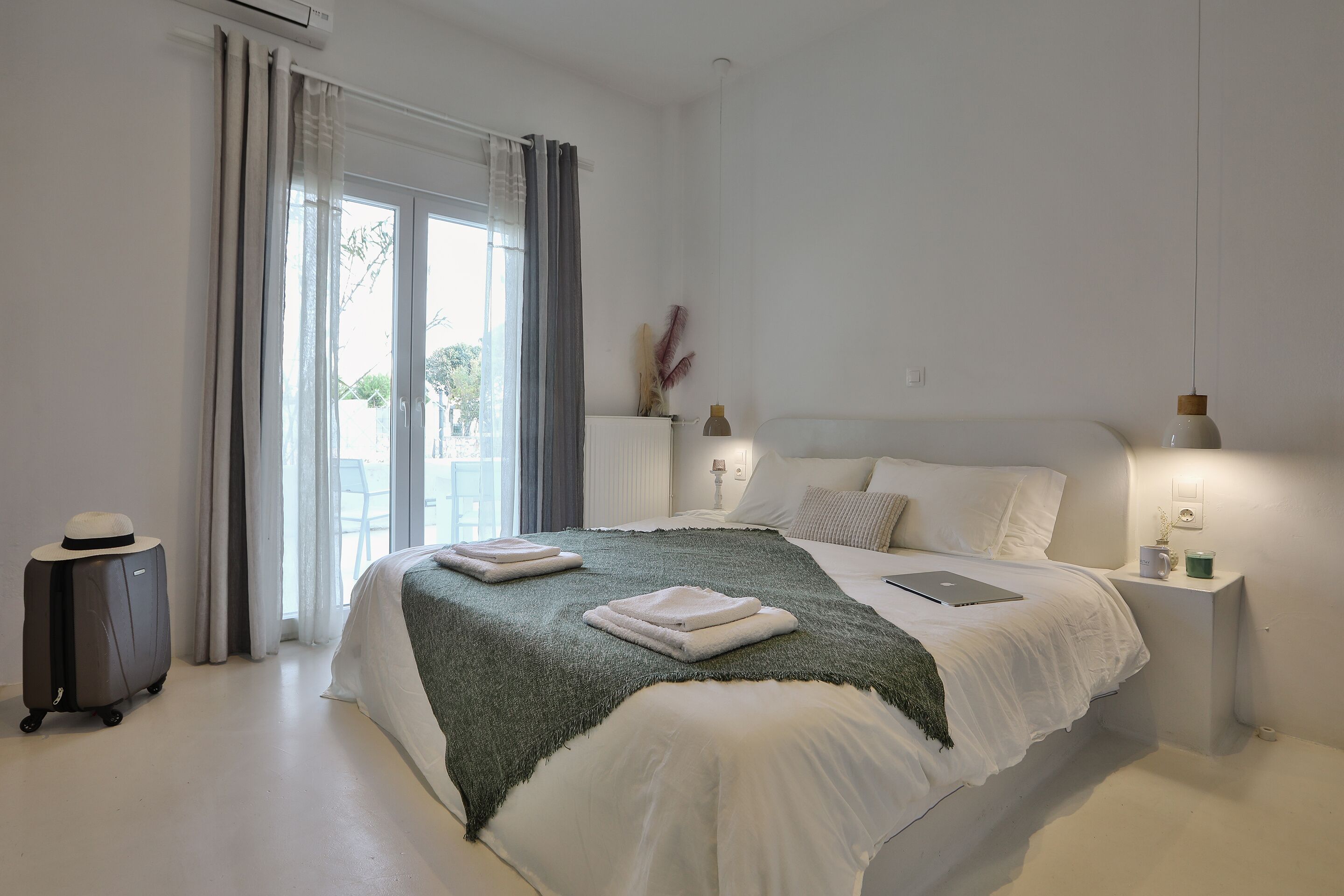 Habitación de hotel con una cama grande, una maleta, una computadora portátil y una mesita de noche con una lámpara y una taza.