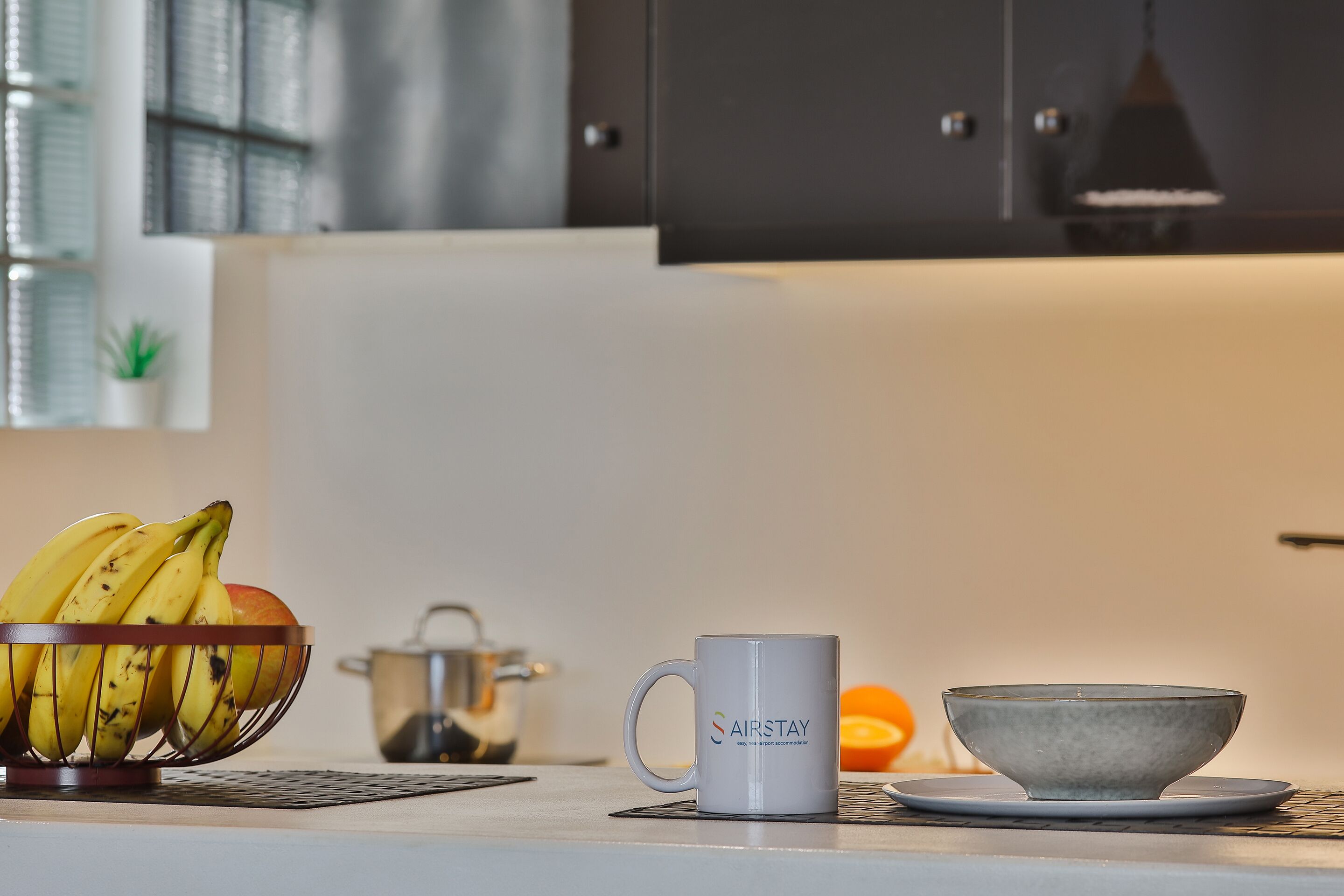 A kitchen counter with a fruit bowl containing bananas and an apple, a white mug with the text "SAIRSTAY," and a gray bowl on a matching saucer.