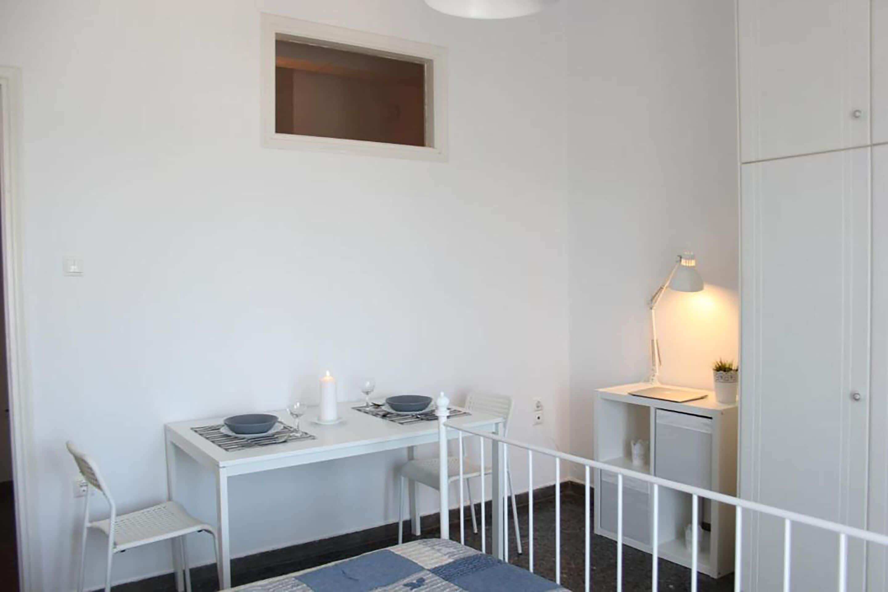 Een kleine, witte kamer met een bureau, twee stoelen, een tafeltje met een kaars, een lamp en een kledingkast.