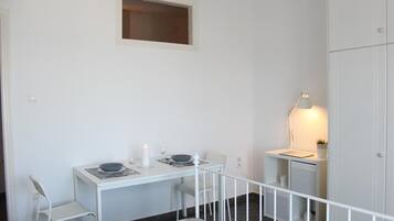Een kleine, witte kamer met een bureau, twee stoelen, een tafeltje met een kaars, een lamp en een kledingkast.