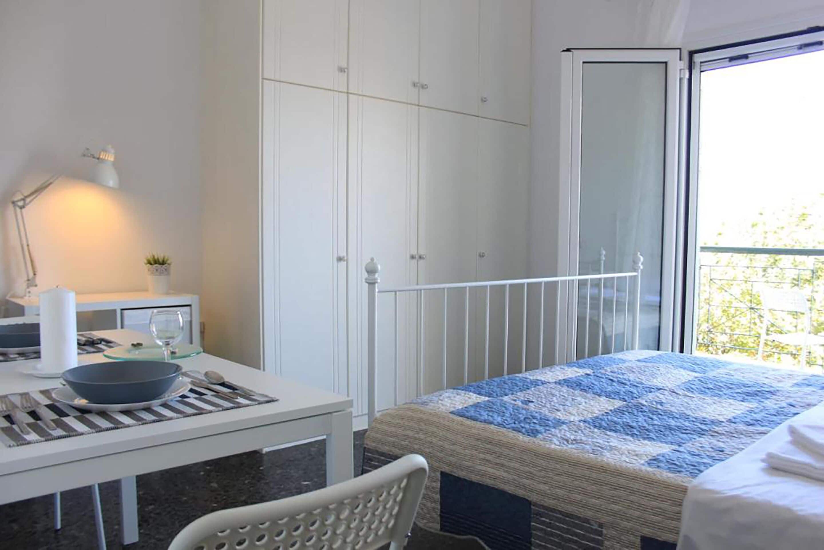 A bedroom with a bed, a desk with a bowl and spoon, a chair, a small table with a lamp, and a wardrobe.