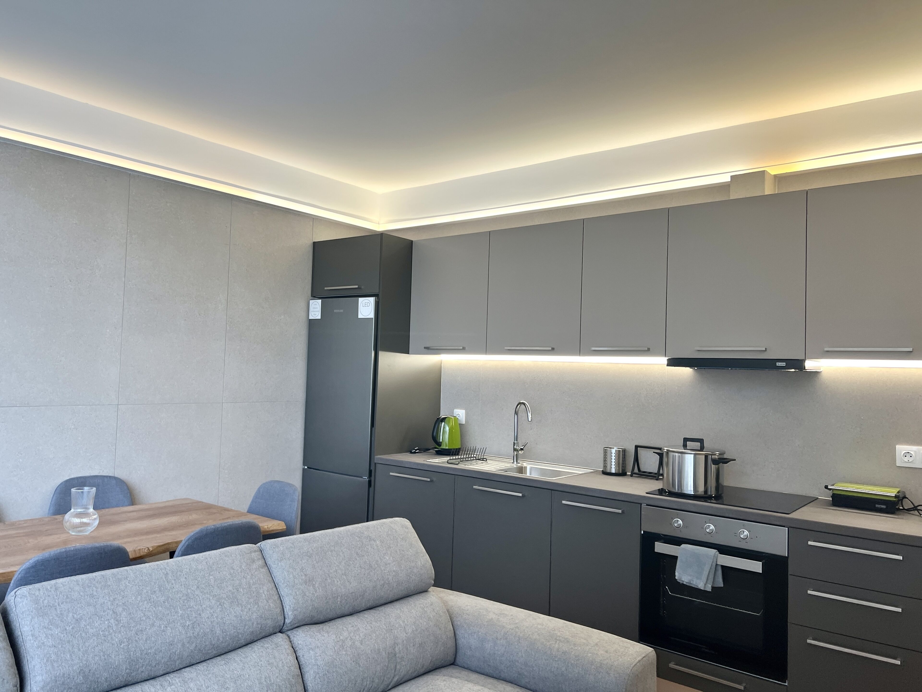 Una cocina moderna con armarios grises, horno empotrado y una zona de comedor con sofá gris y mesa de madera.