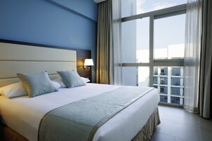 Habitación de hotel con una cama grande, una mesita de noche con lámpara y vistas a edificios a través de la ventana.
