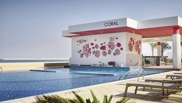 Zona de piscina con vista despejada al mar, un edificio identificado como "CORAL" y sillas de descanso.