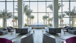 A modern lounge area with large windows, grey sofas, small tables, and a view of palm trees and a distant landmark.