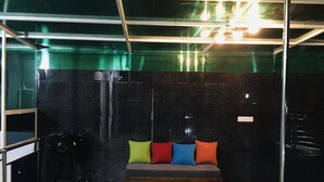 A room with a wooden floor, a black marble wall, a green glass partition, and a wooden bench with colorful cushions. - Grand Star Guest House (Batticaloa)