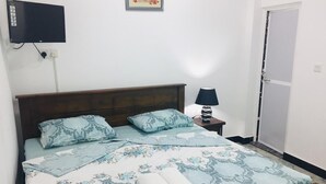 A neatly made bed with floral bedding, a bedside lamp, and a TV mounted on the wall. - Grand Star Guest House (Batticaloa)