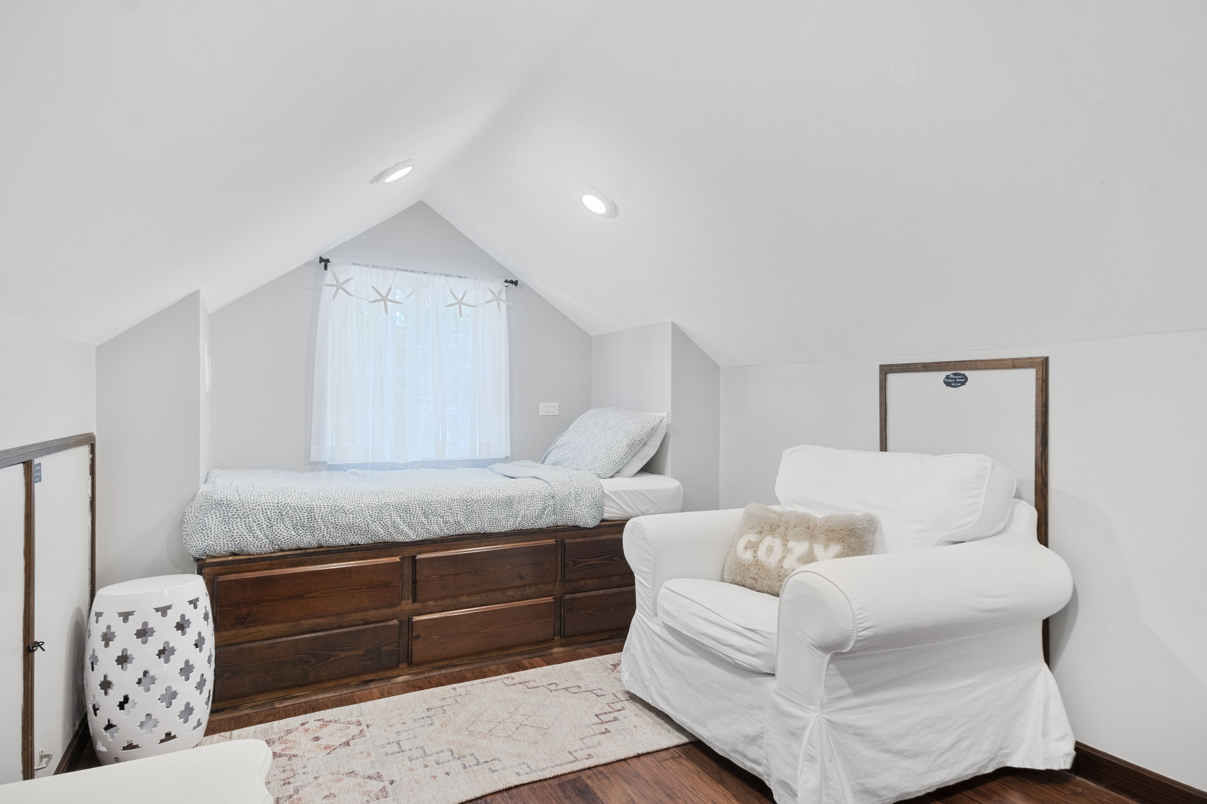 A bedroom with a sloped ceiling, a bed with a white and gray patterned bedspread, a white armchair with a 'COZY' pillow, a wooden dresser, and a small white stool with a geometric pattern.