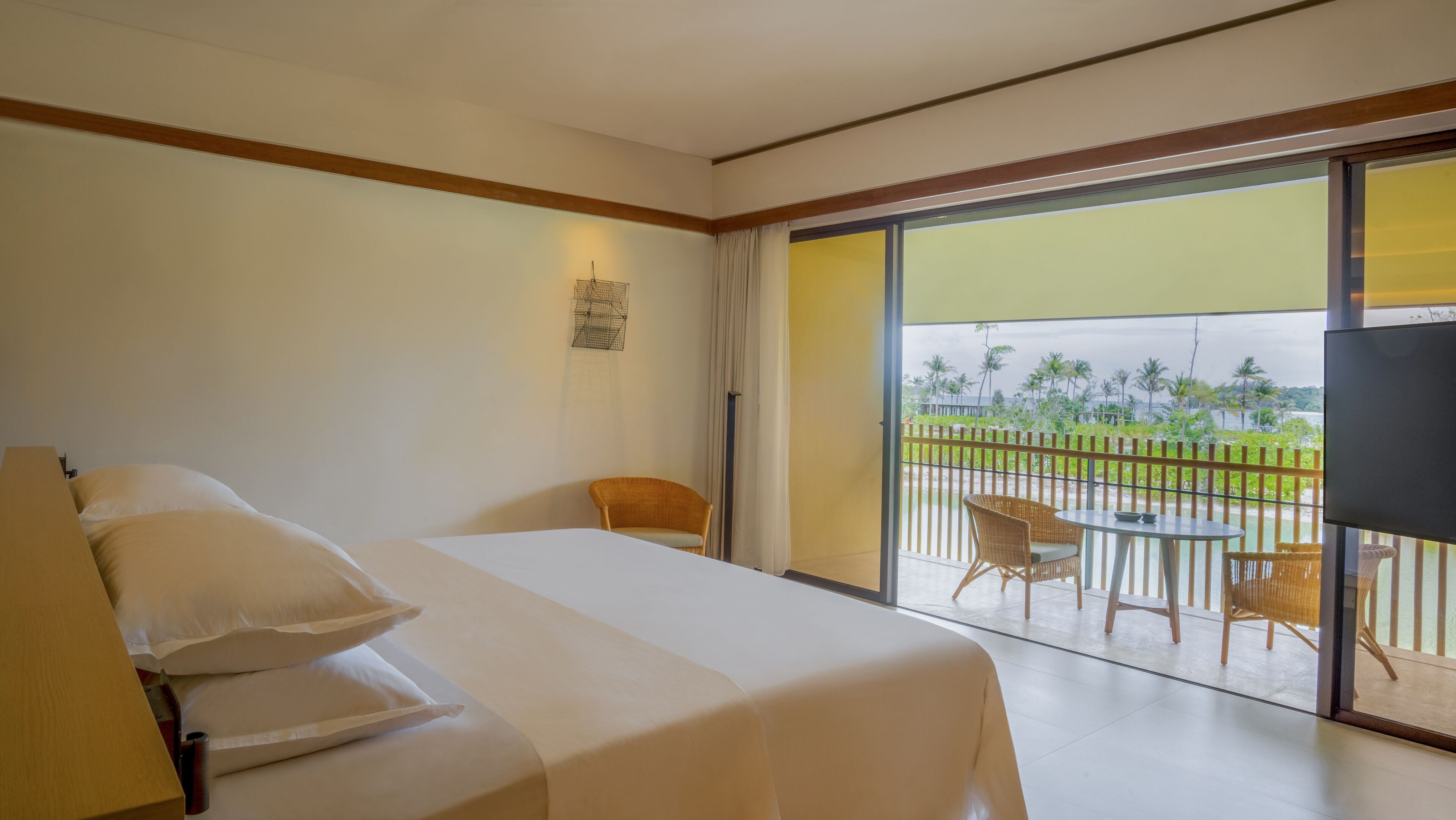Quarto de hotel com cama, cadeira, mesa pequena e varanda com vista para palmeiras e praia.