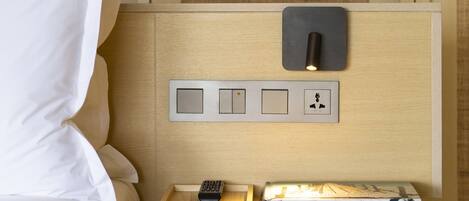 A neatly made bed with white linens, a bedside table with a remote control, a book, and a wall-mounted light switch.