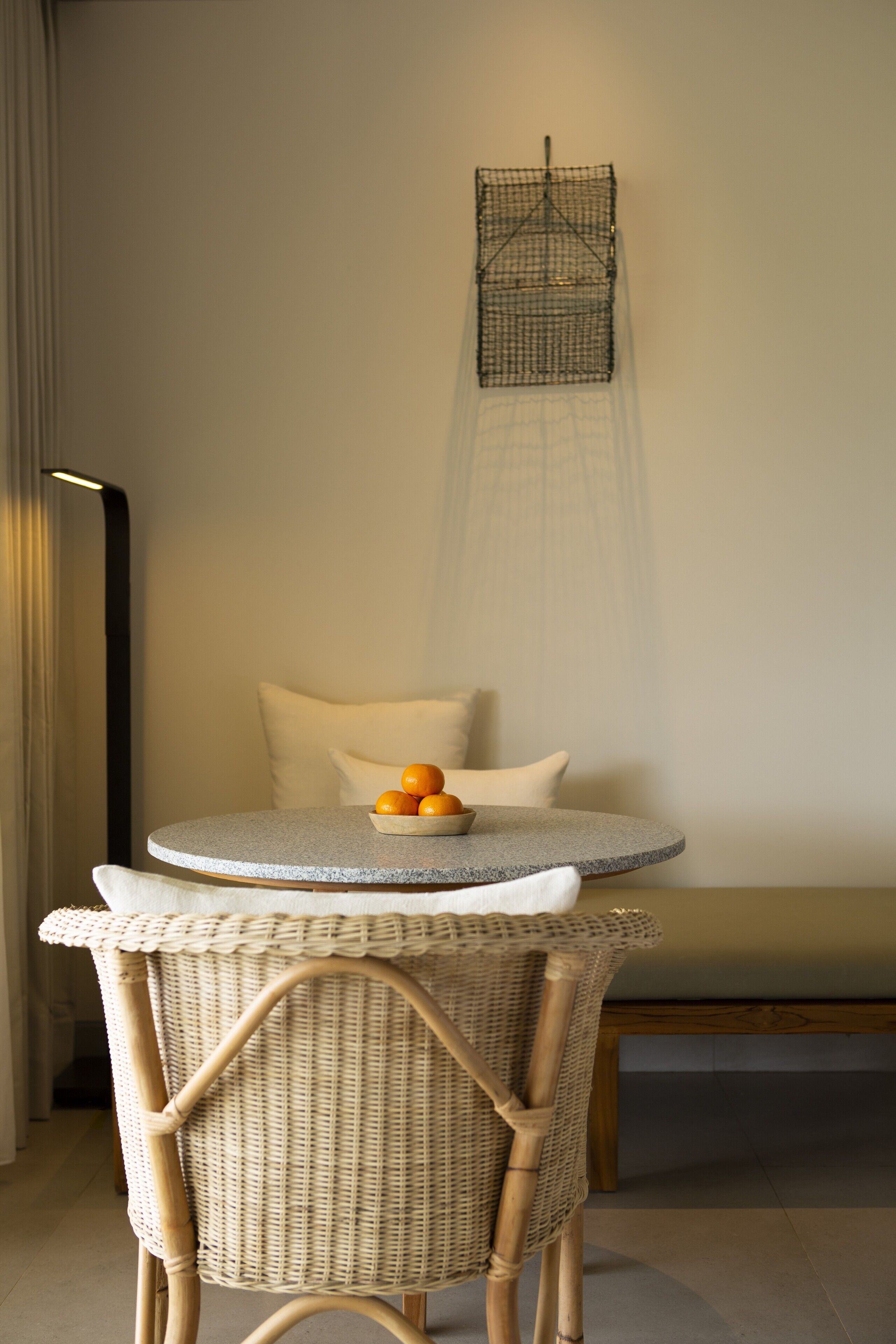 Una mesa redonda con una silla de mimbre, un cuenco de naranjas y un aplique de pared.