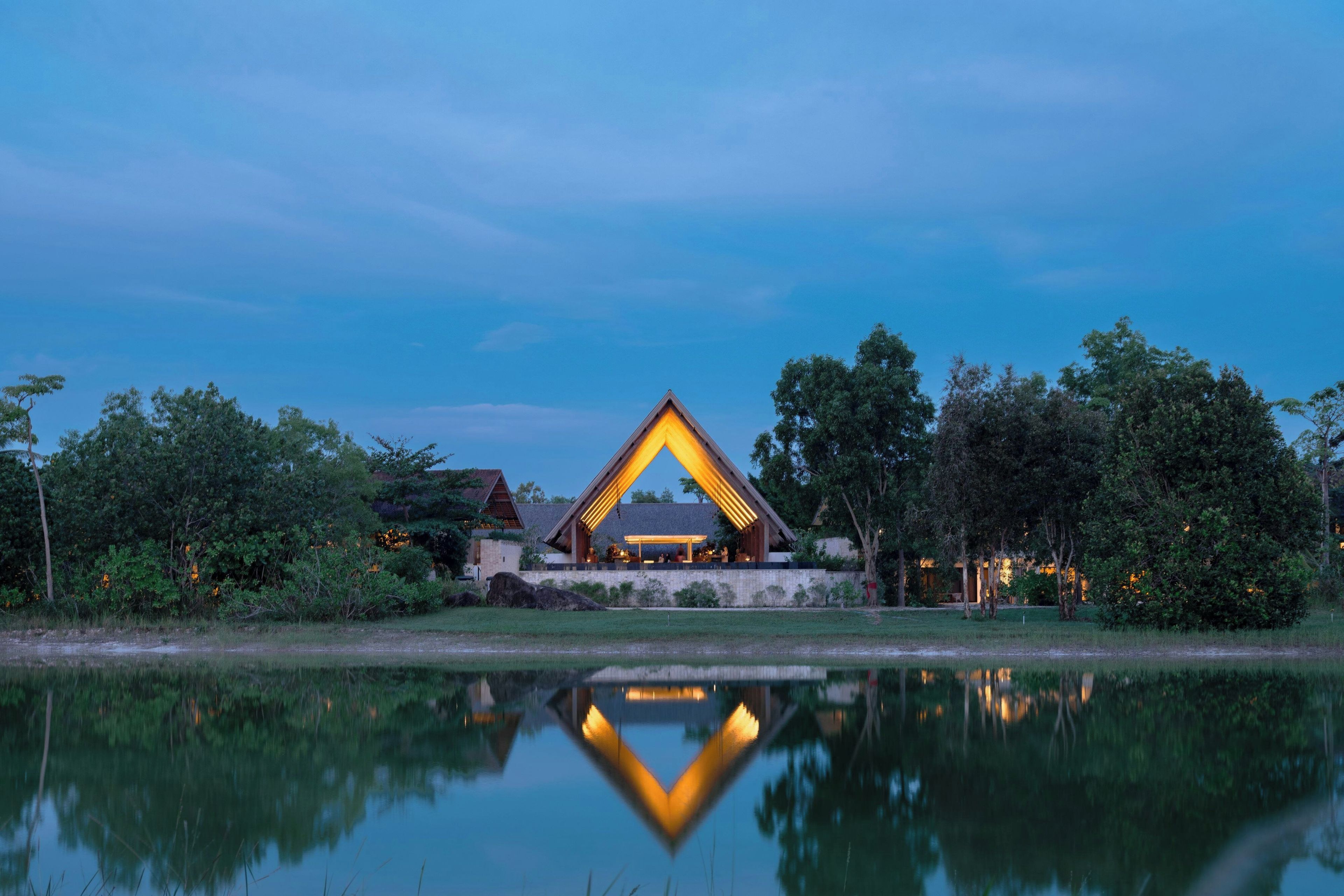 A modern building with a triangular roof structure, surrounded by trees and a body of water reflecting the structure.