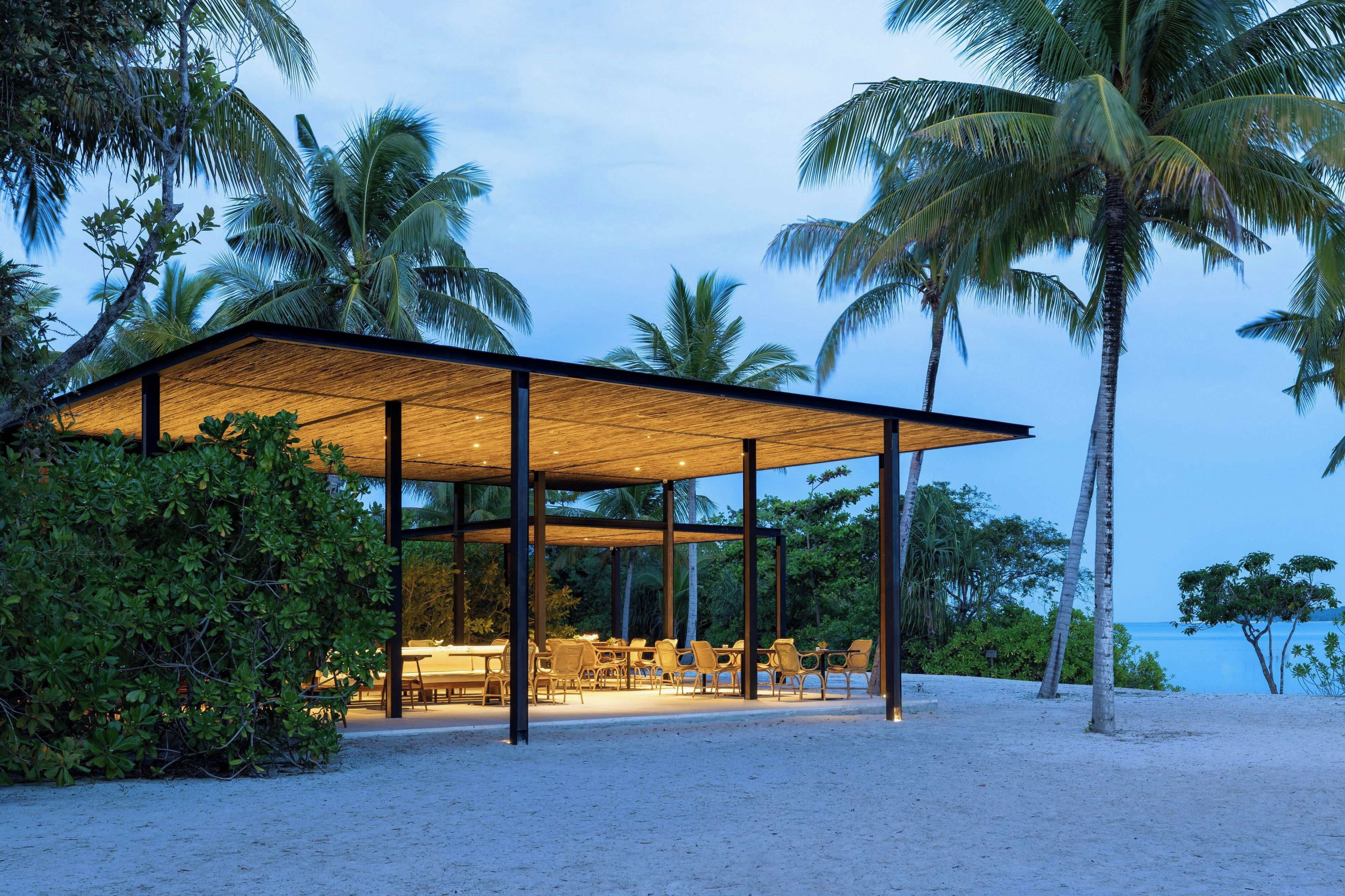 A beachside structure with a glass enclosure, outdoor seating, and palm trees.