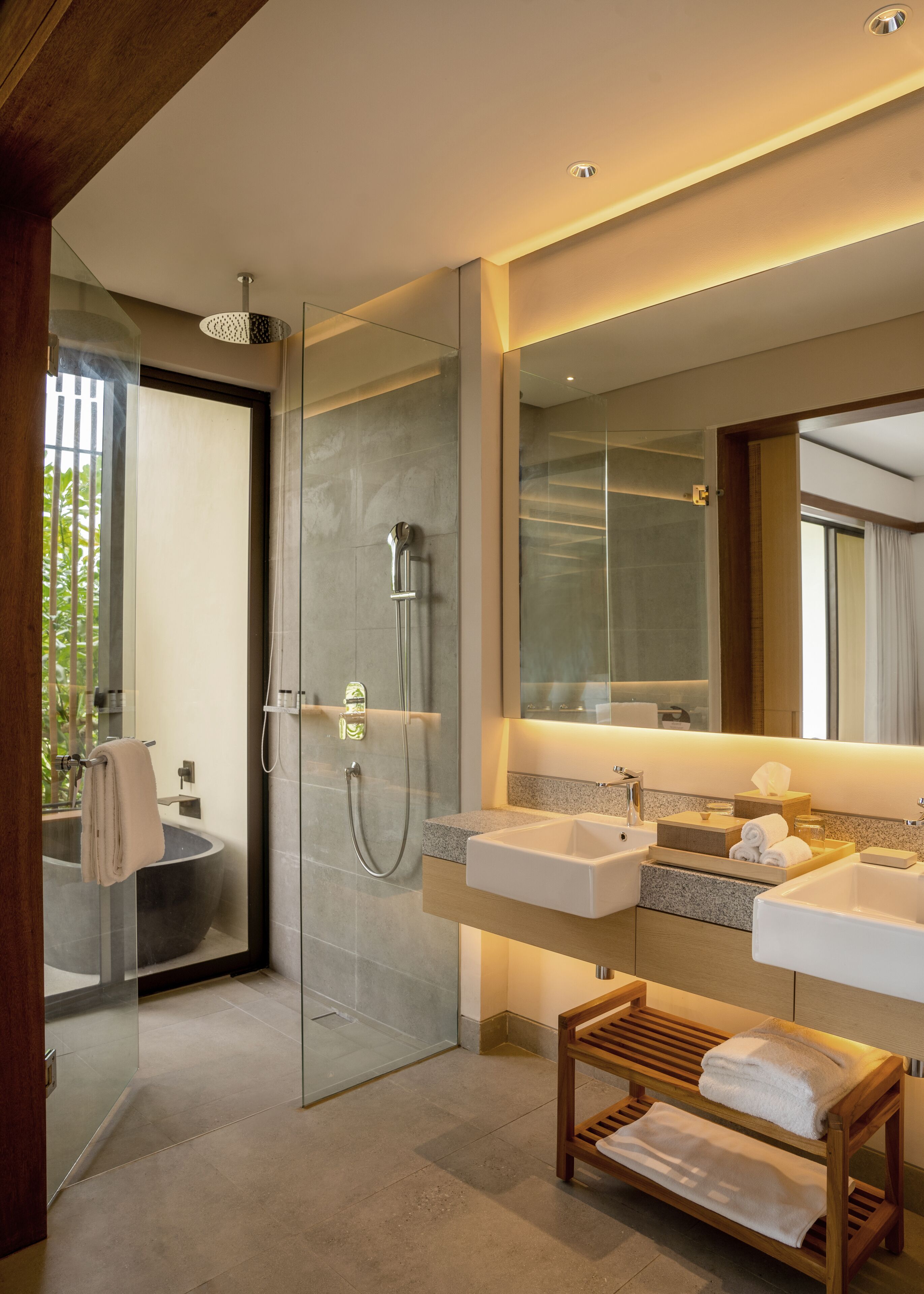 Un baño moderno con cabina de ducha de vidrio, dos lavamanos, un espejo grande y un banco de madera con toallas.