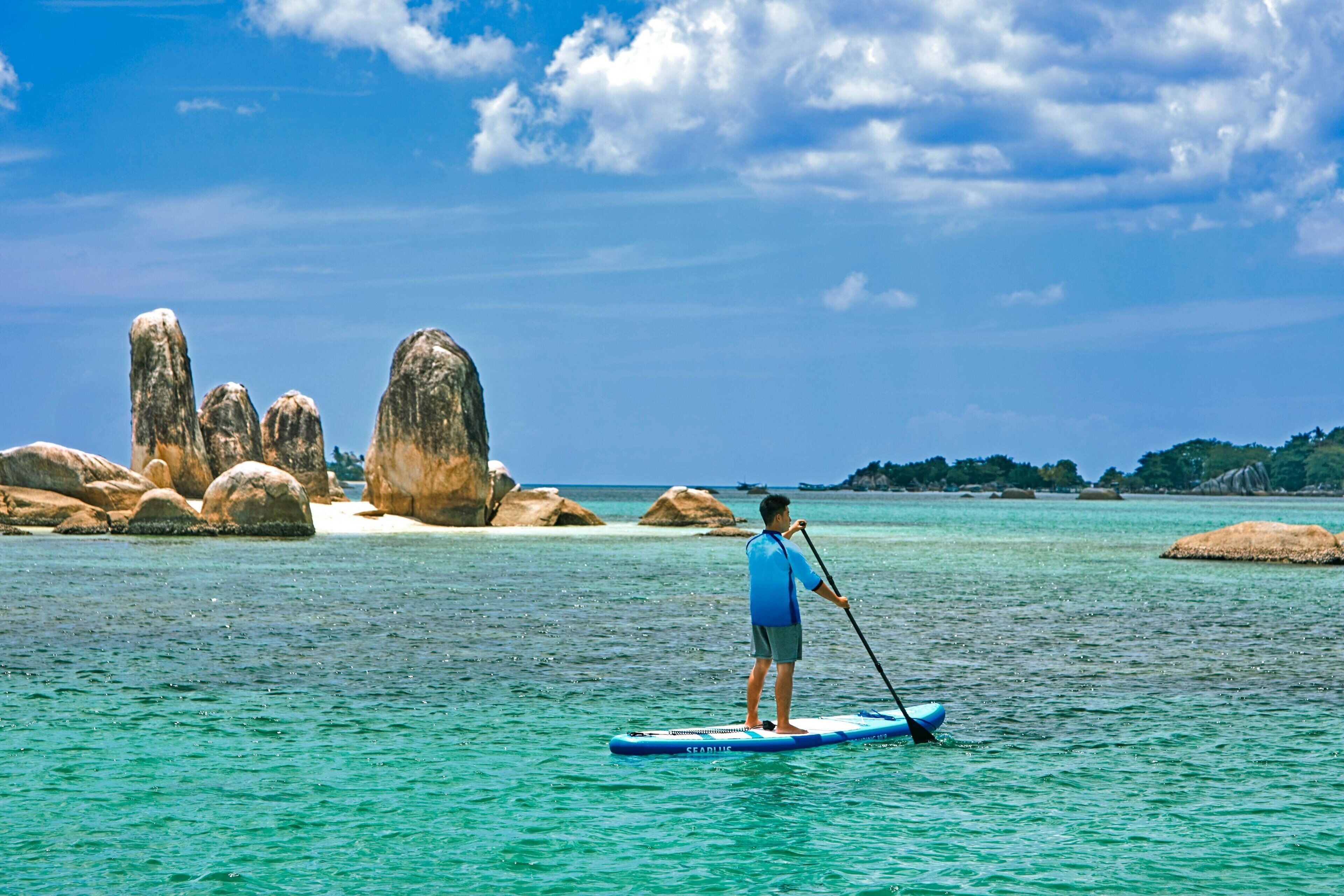 Una persona practicando paddleboard en un mar tranquilo con grandes rocas y cielos despejados.