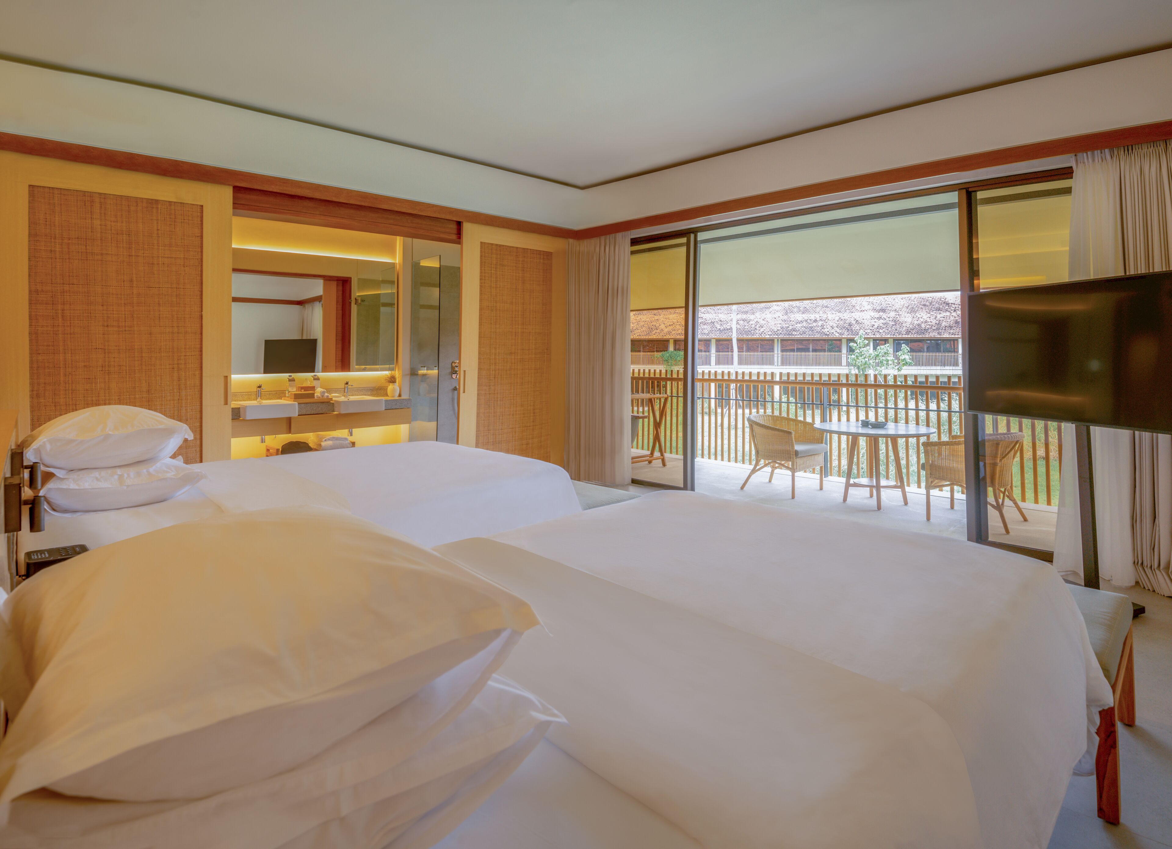 A hotel room with two beds, a balcony with a table and chairs, a TV, and a bathroom with a mirror and sink.
