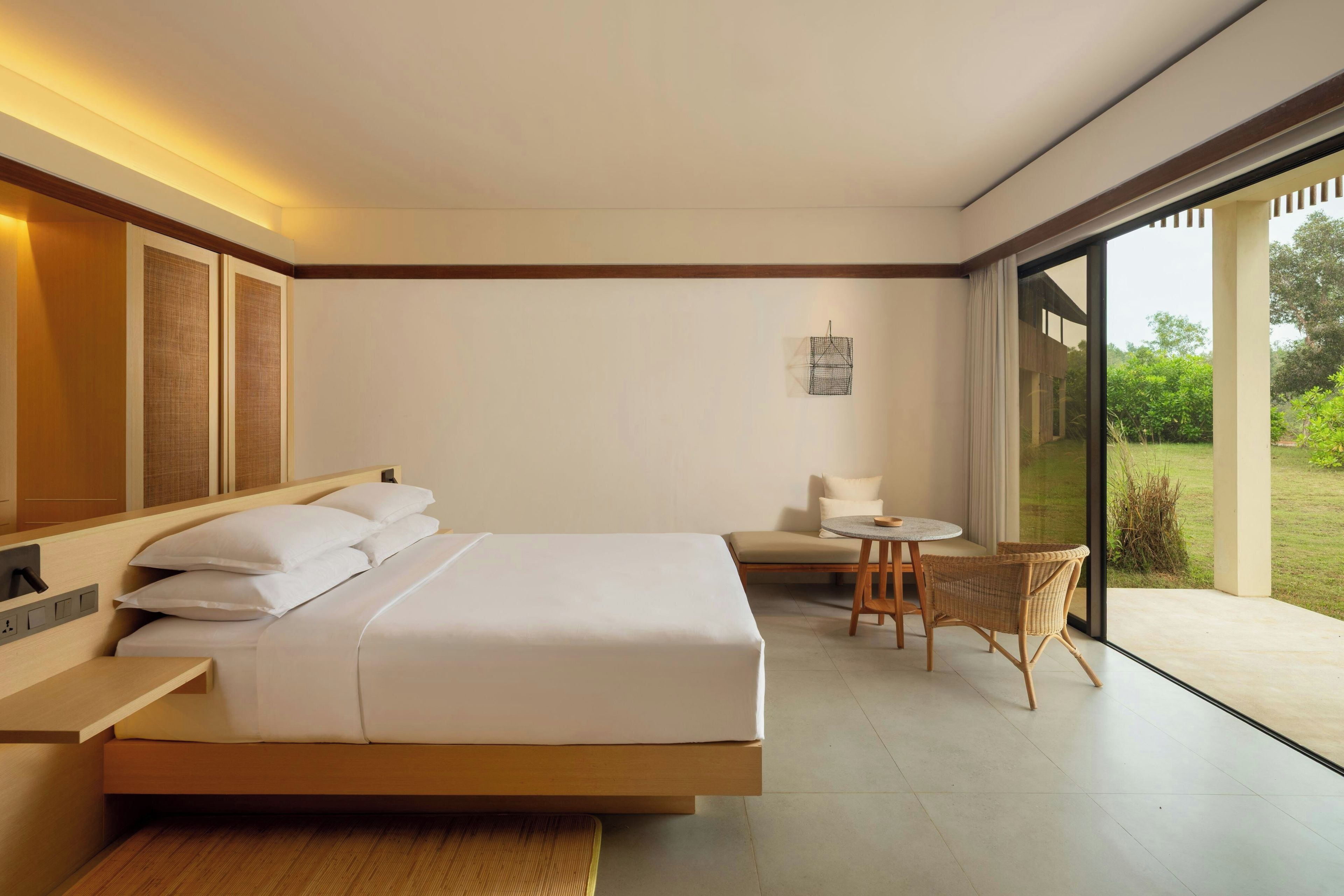 A modern hotel room with a large bed, a small table, and a chair. There is a large window providing a view of the outdoors.