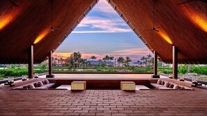 A covered outdoor seating area with a view of a sunset and palm trees. - Sheraton Belitung Resort (Tanjung Pandan)