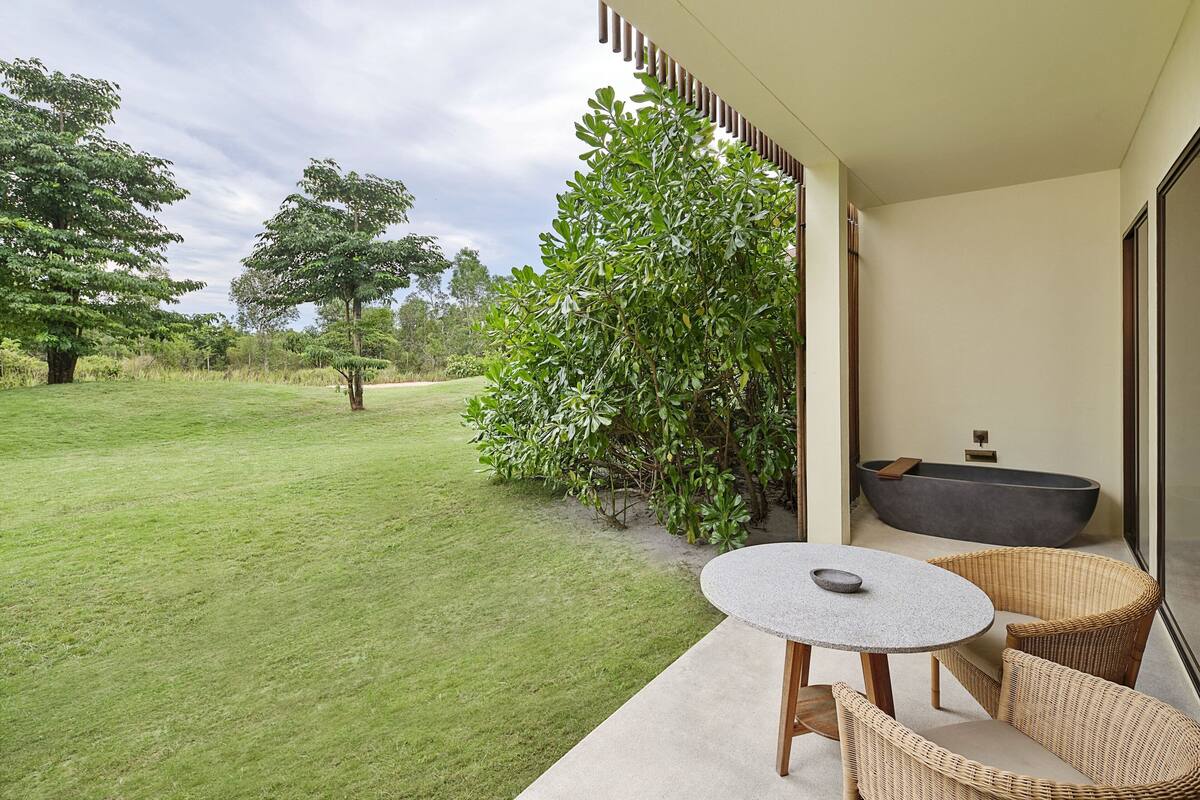 A patio with a round table and wicker chairs, a bathtub, and a view of a grassy area with trees.