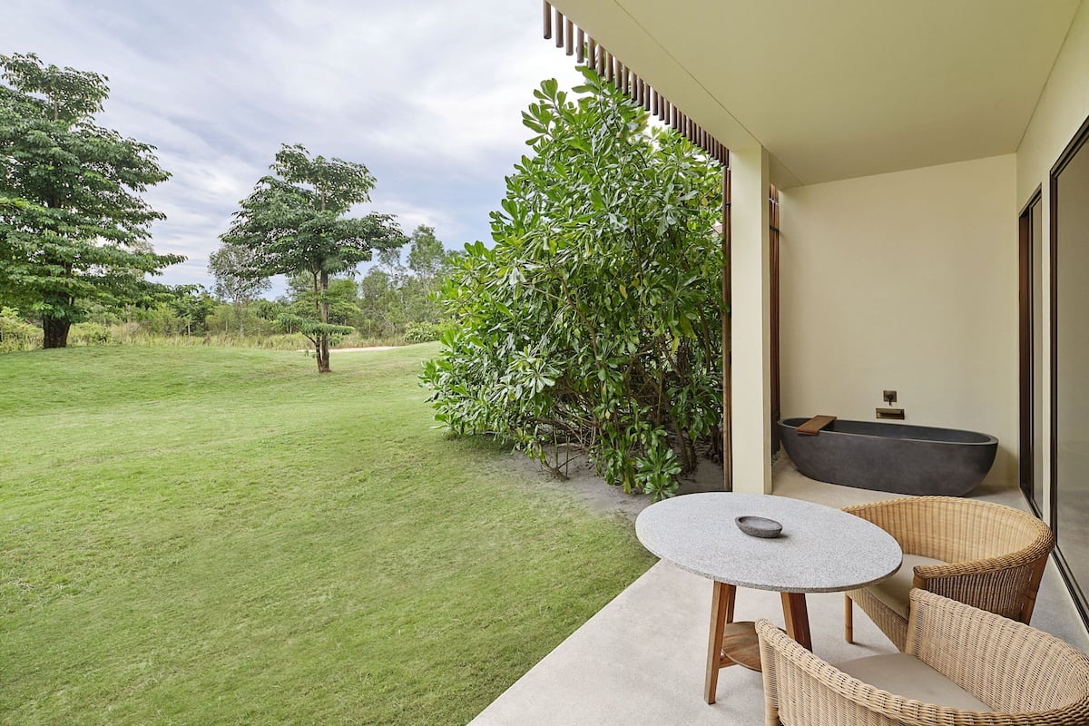 A patio with a round table and wicker chairs, a bathtub, and a view of a grassy area with trees.