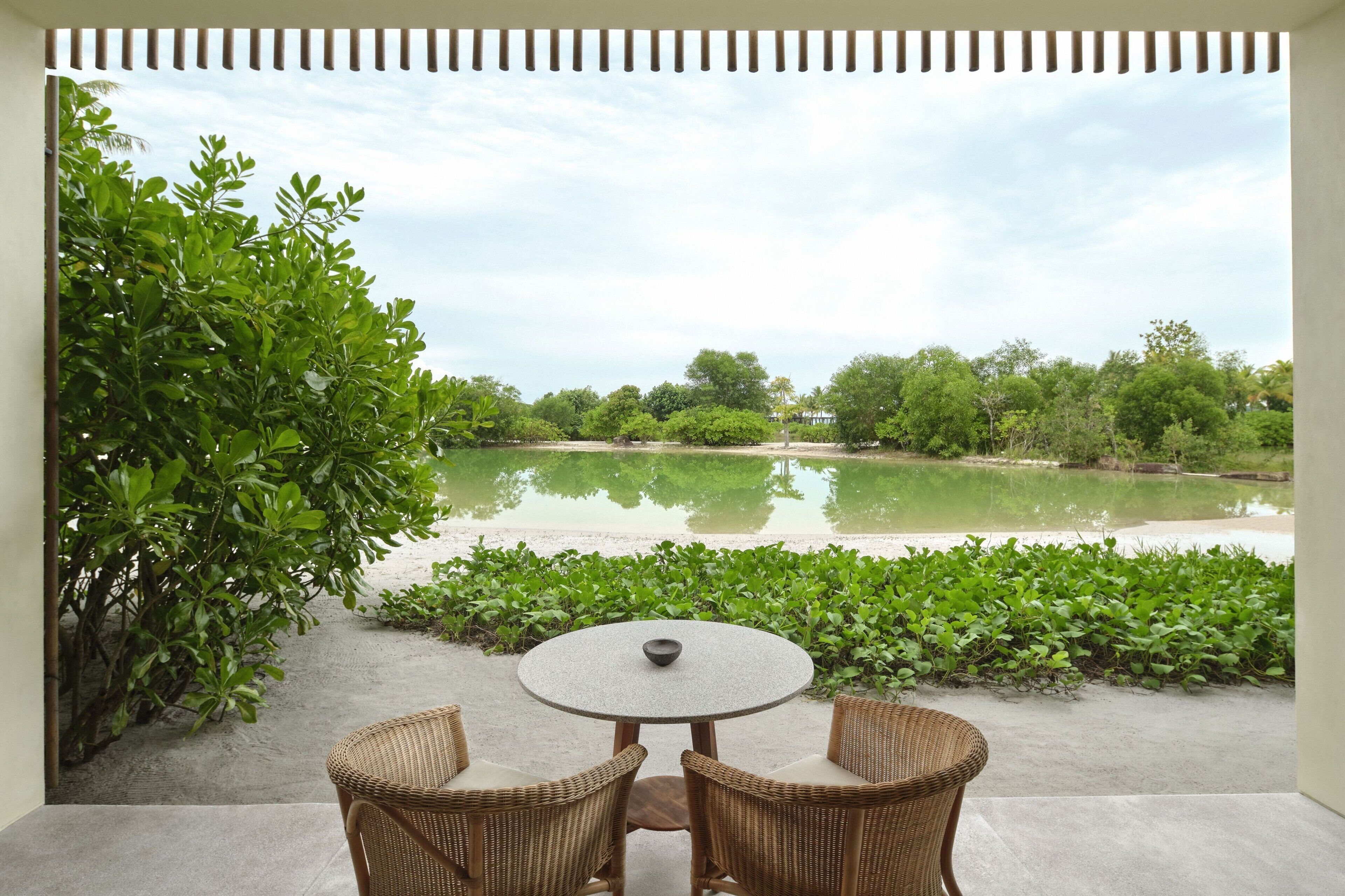 An outdoor seating area with a round table and two wicker chairs, overlooking a calm body of water and lush greenery.