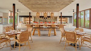 A spacious dining area with a large wooden table and wicker chairs, a kitchen in the background, and large windows offering a view of the outdoors. - Sheraton Belitung Resort (Tanjung Pandan)