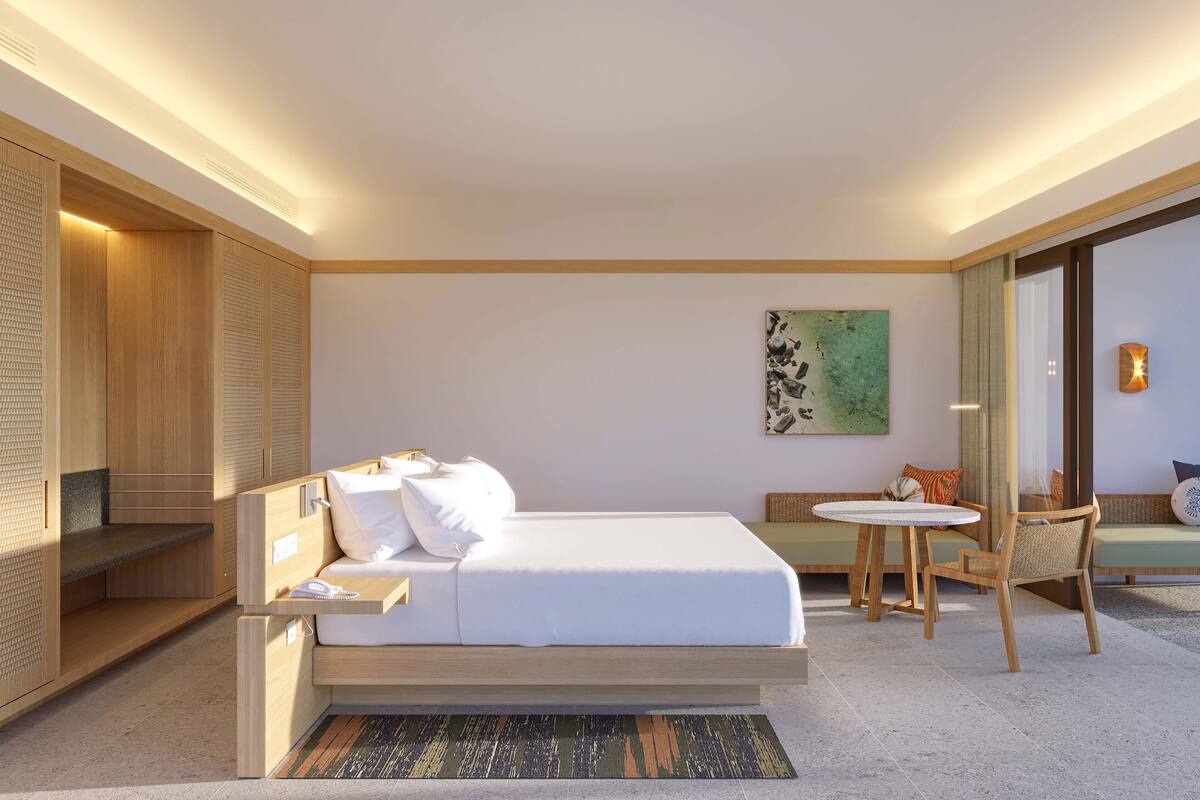 A modern hotel room with a bed, bedside table, seating area, and a view of the outdoors.