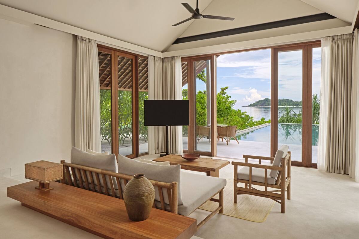 A spacious room with a large window offering a view of a body of water and greenery. There is a flat-screen TV on a wooden table, a sofa, and a chair.