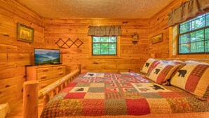 A cozy log cabin room with a wooden bed, a television on a dresser, and a window with a view of trees. - Secret Hideaway #195 by Aunt Bug's Cabin Rentals (Sevierville)