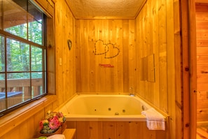 A wooden cabin with a large bathtub, a window with a view of trees, and a heart-shaped decoration on the wall.