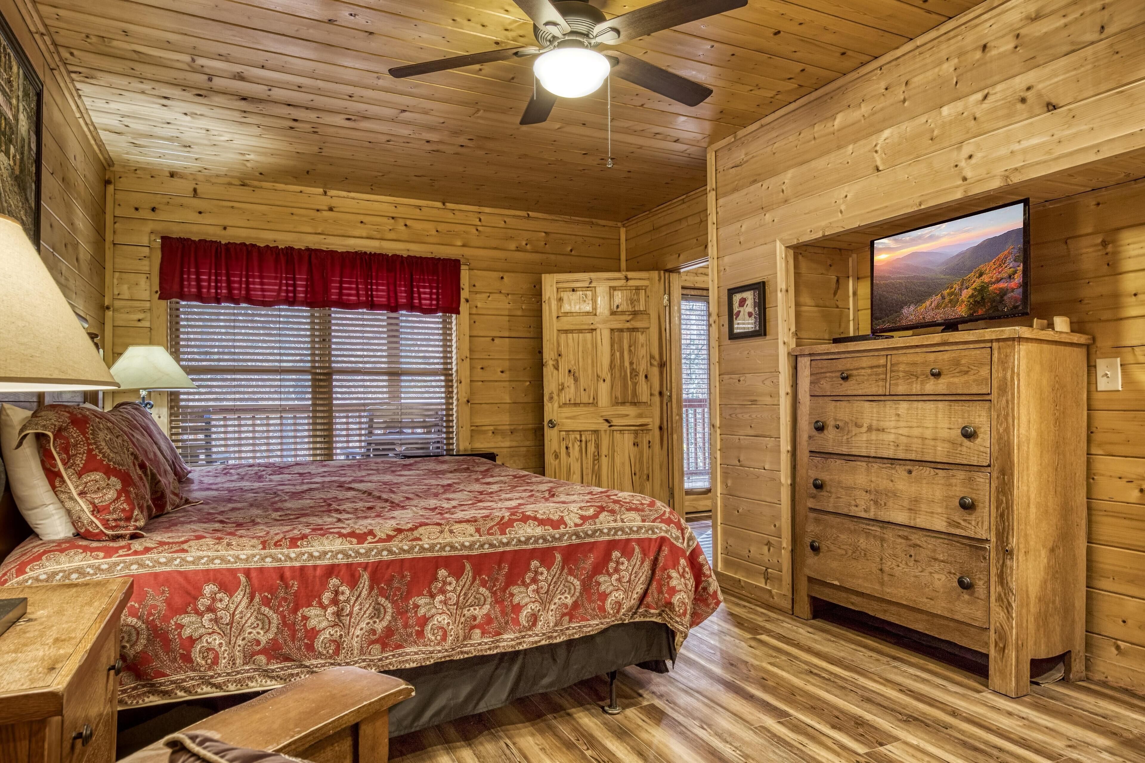 Ein Zimmer in einer Holzhütte mit einem Bett, einem Schminktisch, einem Deckenventilator und einem Fernseher auf einem Holzkasten.