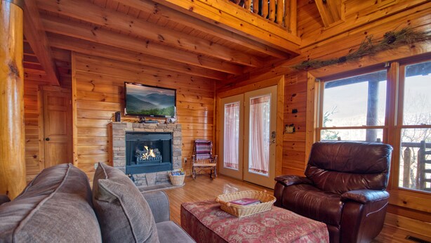 A cozy log cabin interior with a fireplace, leather armchair, and a large window. - Poolside Retreat #221 by Aunt Bug's Cabin Rentals (Sevierville)