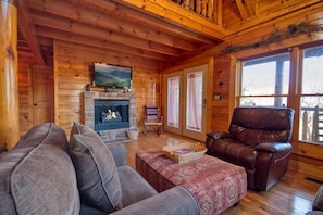 Acogedor interior de una cabaña de madera con chimenea, un sillón de cuero y un ventanal grande.