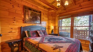 A wooden cabin bedroom with a bed, a nightstand, a lamp, and a window with a view of trees. - Poolside Retreat #221 by Aunt Bug's Cabin Rentals (Sevierville)