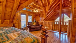 A cozy log cabin bedroom with a large bed, a leather sofa, and a balcony with a wooden railing. - Poolside Retreat #221 by Aunt Bug's Cabin Rentals (Sevierville)