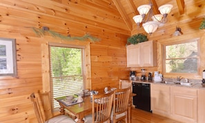L’intérieur d’un chalet en bois comprend une table à manger, des chaises, un coin cuisine avec des placards et des appareils électroménagers, ainsi qu’une fenêtre équipée de stores.