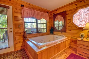 A wooden cabin bathroom with a large bathtub, a window with blinds, a wooden vanity, and a decorative mirror. - Smoky Mountain Getaway #435 (Sevierville)