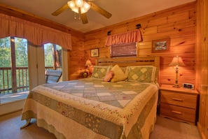 A cozy bedroom with a wooden cabin aesthetic, featuring a large bed with a quilt, a nightstand with a lamp, and a window with blinds. - Smoky Mountain Getaway #435 (Sevierville)