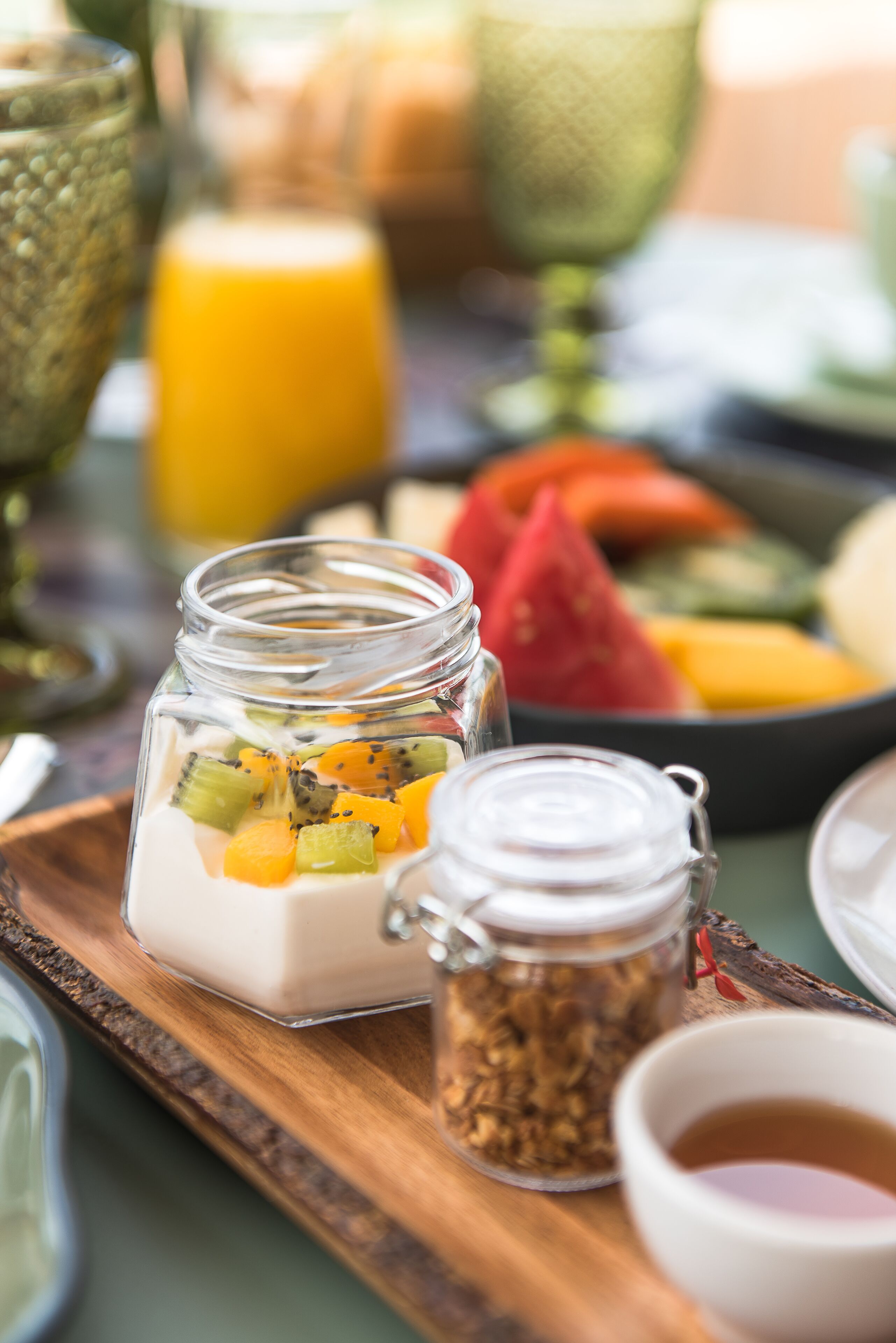 A wooden tray with a glass jar containing yogurt and fruit, a small jar of granola, and a cup of tea.
